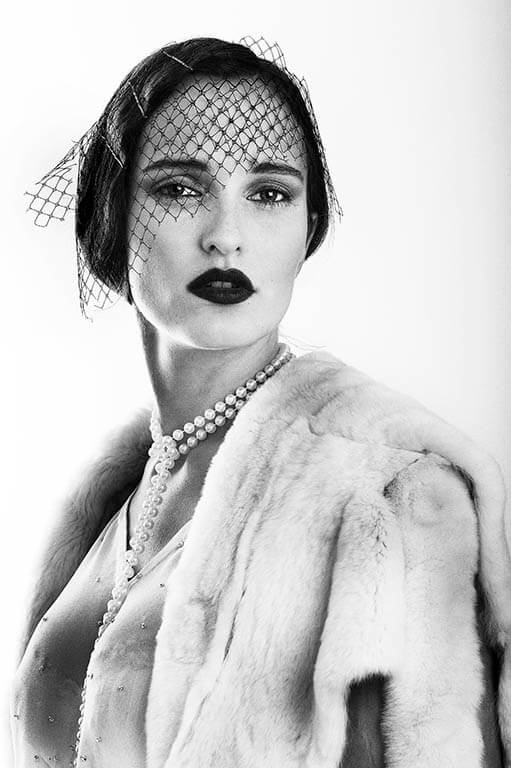 Elegant woman in a vintage-style portrait with a veil, pearls, and fur coat, exuding sophisticated mystery.