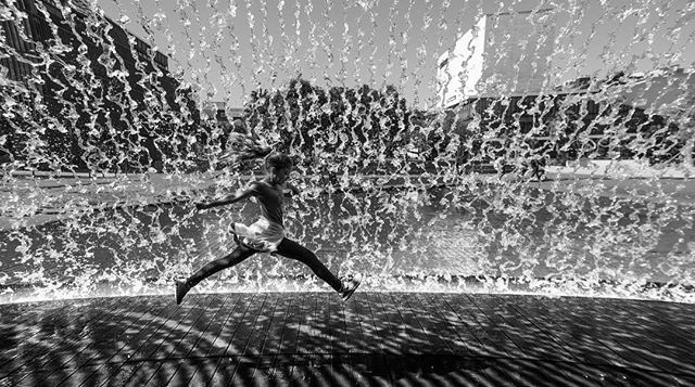 A woman jumps happily in front of a water fountain in an urban setting, a joyful moment captured in monochrome.
