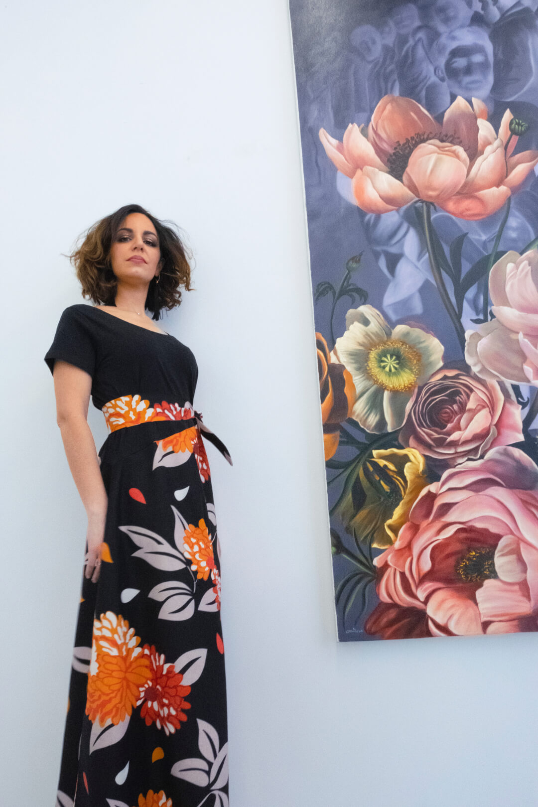 A woman in a floral dress poses beside a floral painting in a modern art gallery.