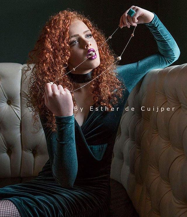 Elegant redhead woman poses with a necklace on a vintage couch in a soft-lit studio, perfect for beauty and fashion campaigns.