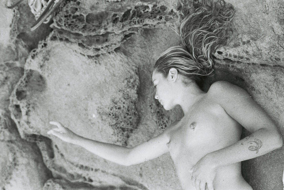 Artistic black and white image of a nude woman lying on rocks by the beach in a moment of serene relaxation.
