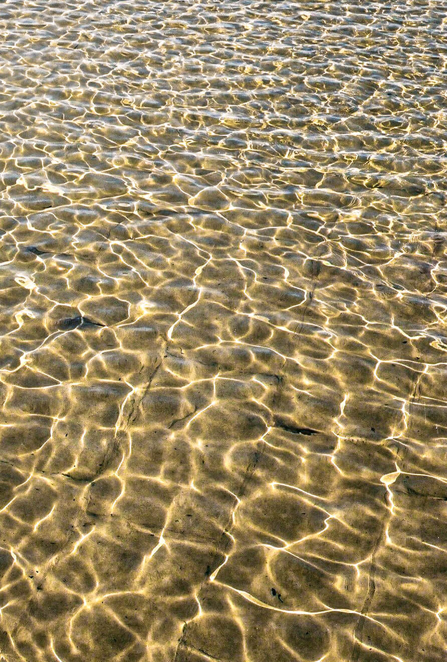 Sunlight ripples across the clear, shallow water creating abstract patterns on the sandy seabed.