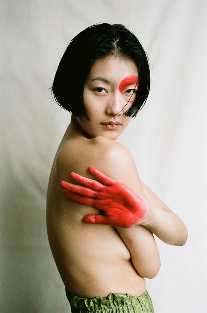 A nude Asian woman with red paint on her face and hand poses in a studio setting.