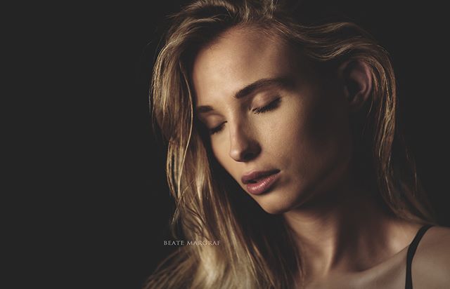 A serene woman with blonde hair is captured in a dreamy portrait against a dark backdrop.