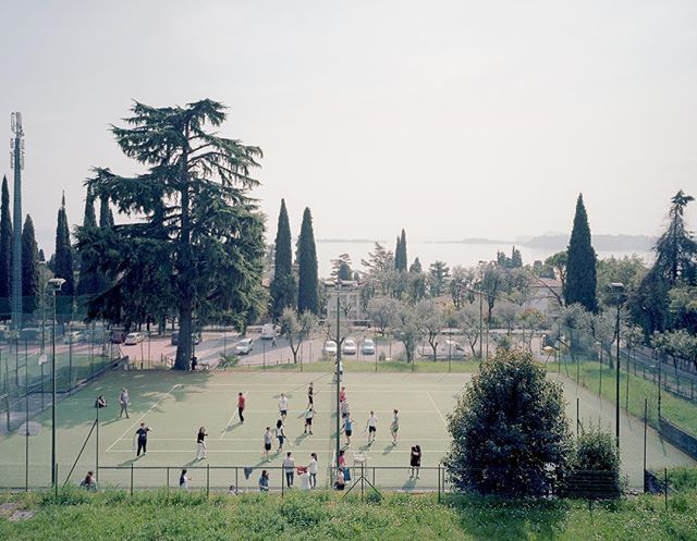 A group of people are playing tennis on an outdoor court surrounded by trees on a bright day.