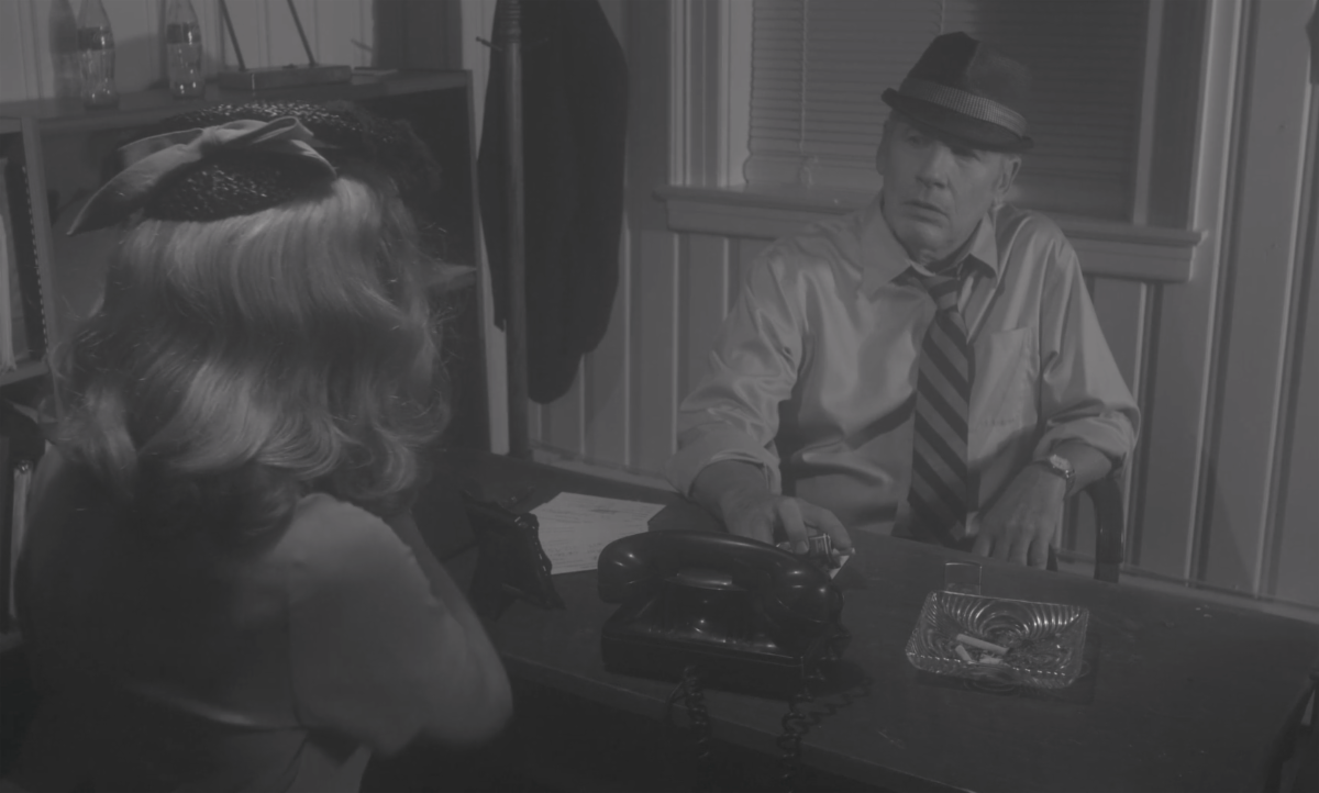 Still from the film Private, starring Sandy Bainum and Steve Blackwood. The black-and-white image shows the back of a blonde woman's head. She is seated across a desk from a man wearing a tie and a fedora.