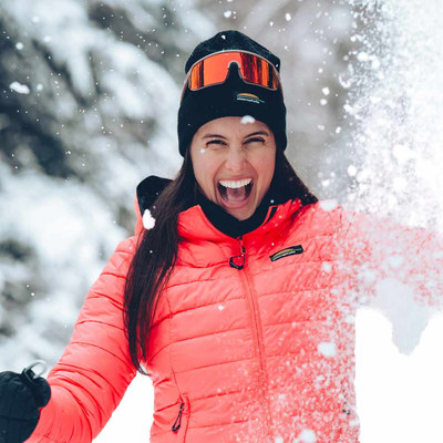 Femme souriante jouant dans la neige, vêtue d’une veste d’hiver Chlorophylle, ambiance hivernale lumineuse.
