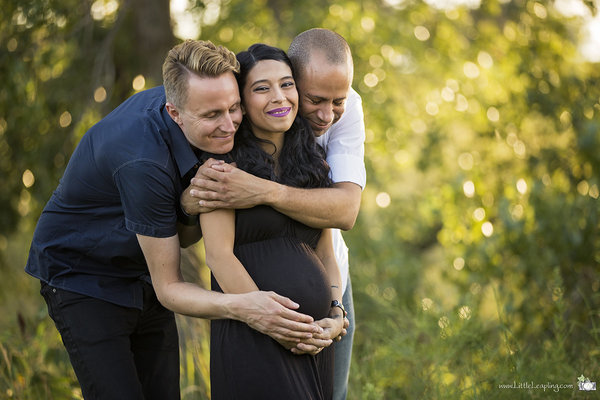 Pregnant woman smiling and cradling belly while being embraced by two happy men.