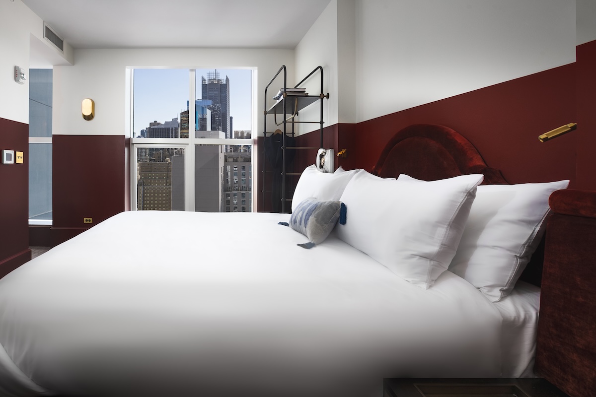 A hotel room with a large bed, city view, and burgundy accent wall.