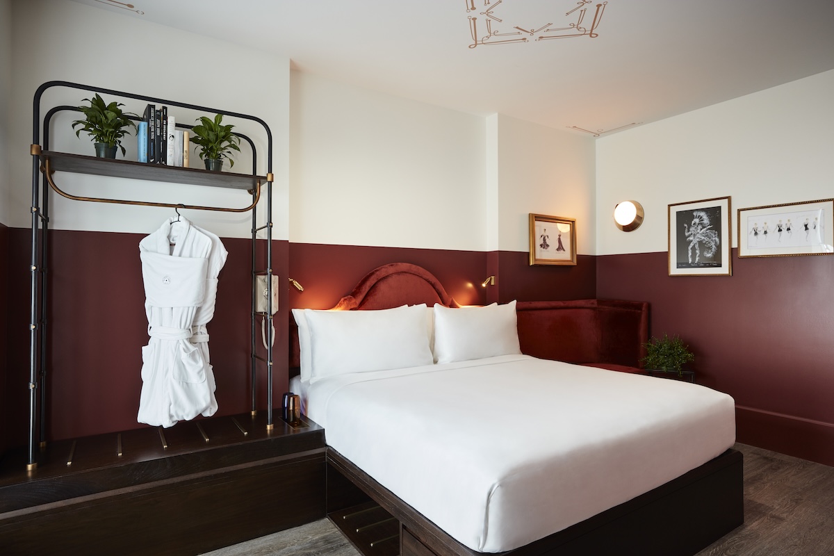 A hotel room with a bed, robe, shelf with plants and books, and framed pictures on the wall.