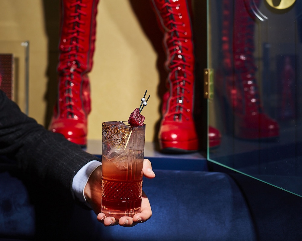 A hand holding a cocktail with red boots in the background.