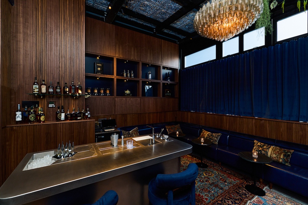 A dimly lit bar with blue leather booths, a stainless steel bar top, and a large chandelier.