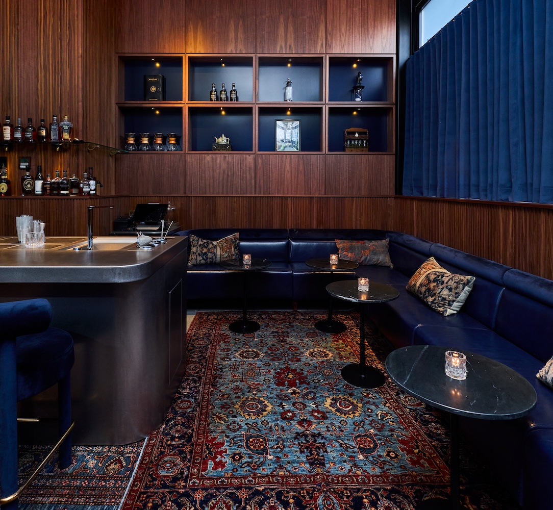 A dimly lit bar with a blue couch, coffee tables, and a bar area with shelves of bottles.