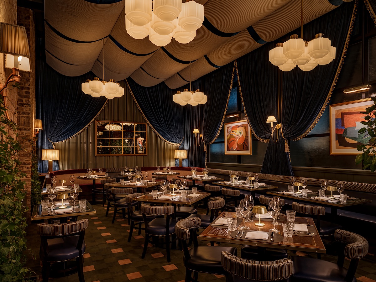 A dining area with tables and chairs, set with glasses and silverware, under a ceiling with hanging lights and draped curtains.