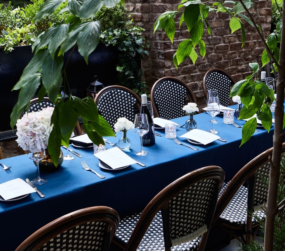 A beautifully set outdoor dining table with a blue tablecloth, white flowers, and wine glasses in a garden setting with a brick wall.