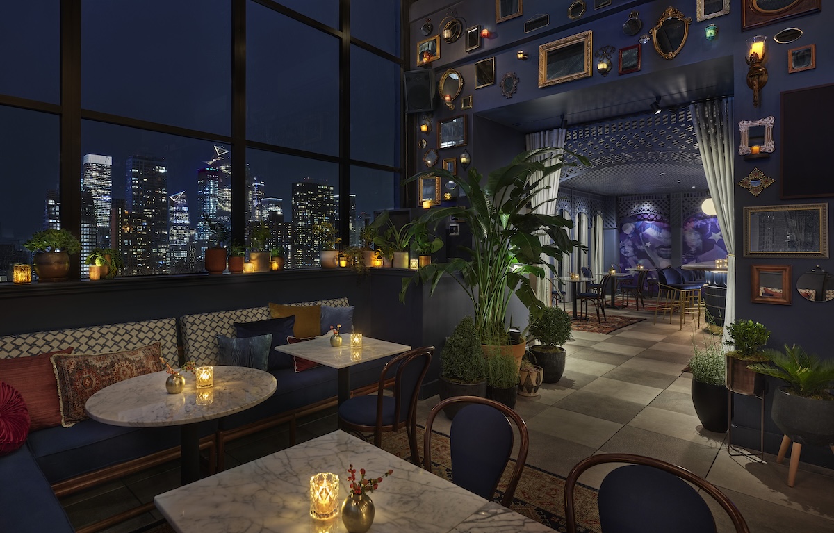 A dimly lit lounge with marble tables, booths, and plants, offering a city view at night.