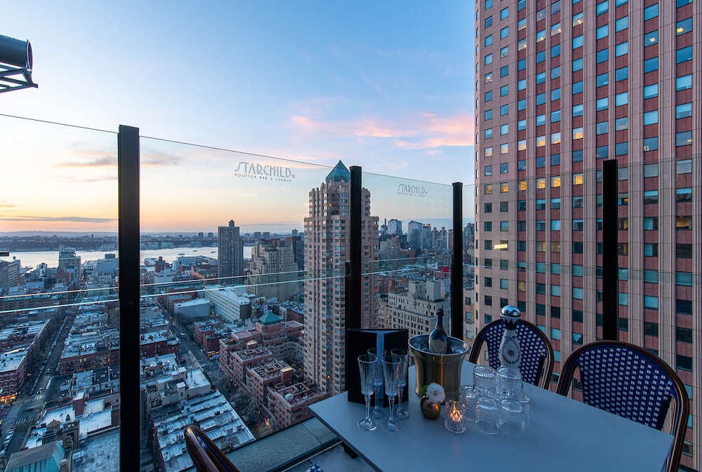 A rooftop bar with a table set for two, overlooking the city skyline at sunset.