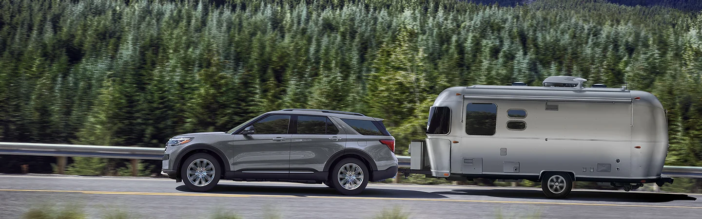 Side view of a gray 2026 Ford Explorer towing a trailer next to a forest