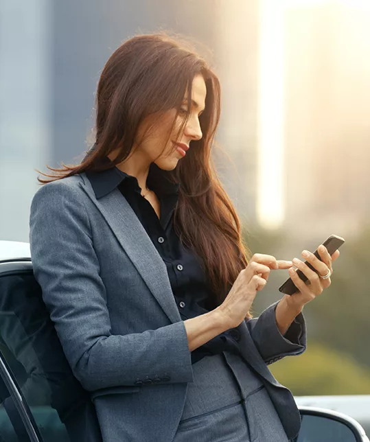 woman dressed in a suit leaning up against a car looking at her phone