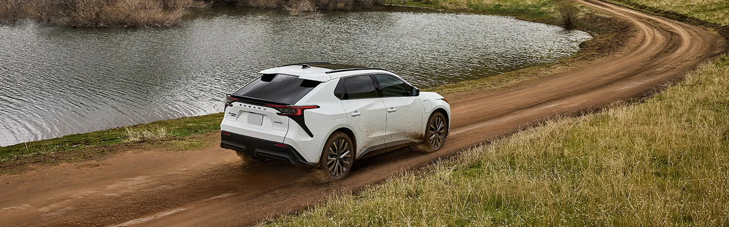 Back side ivew of a white 2026 Subaru Solterra driving on a dirt road by a lake