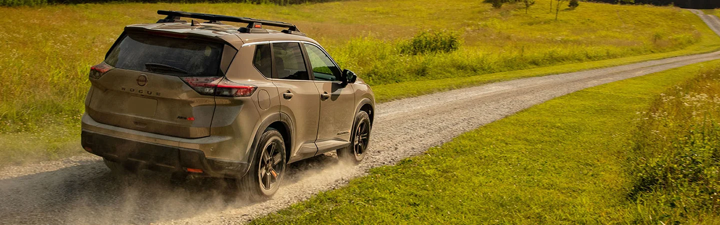 Back side view of a tan 2026 Nissan Rogue driving on a dirt road