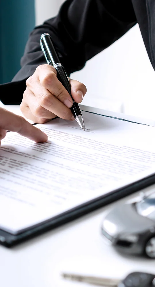 two hands in view one holding a pen working on a desk with papers to sign