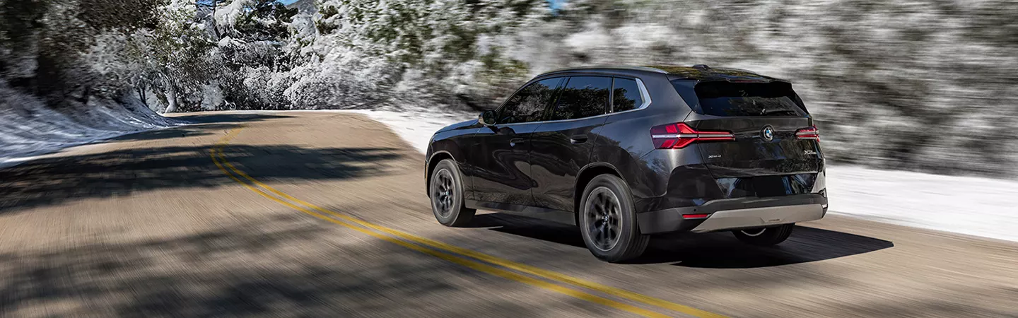 Rear view of 2026 BMW X3 30 xDrive driving on a country road with snow in the background