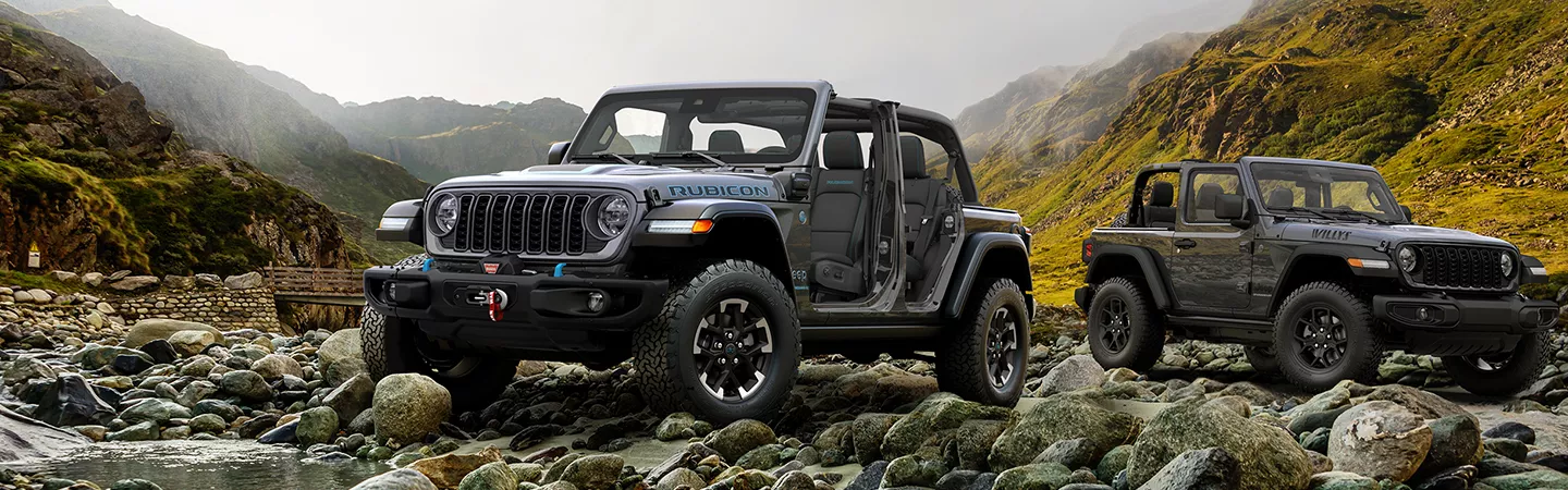 Two 2026 Jeep Wranglers parked on rocks near a creek with mountains in the background