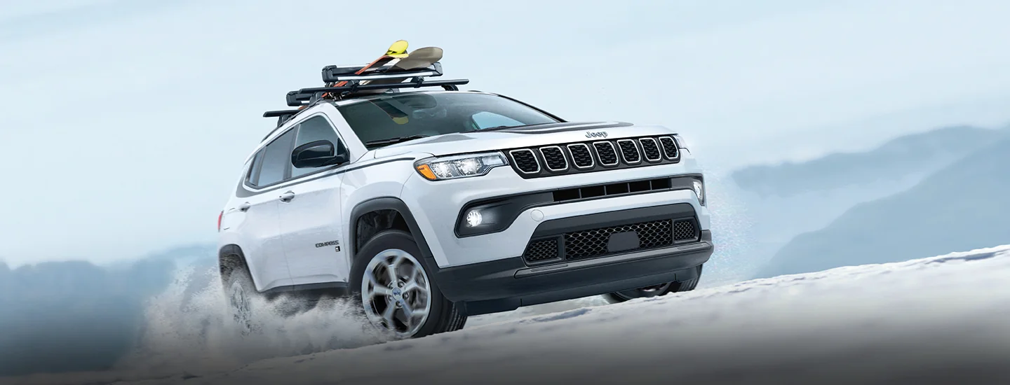 a white 2026 Jeep Compass Limited driving in snow with ski gear mounted on roof rack on top