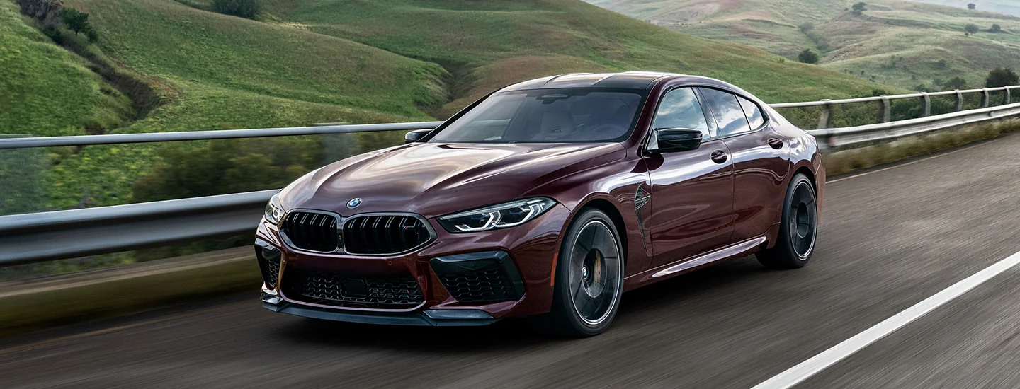 Red 2025 BMW M8 Coupe driving on a road next to some hill sides