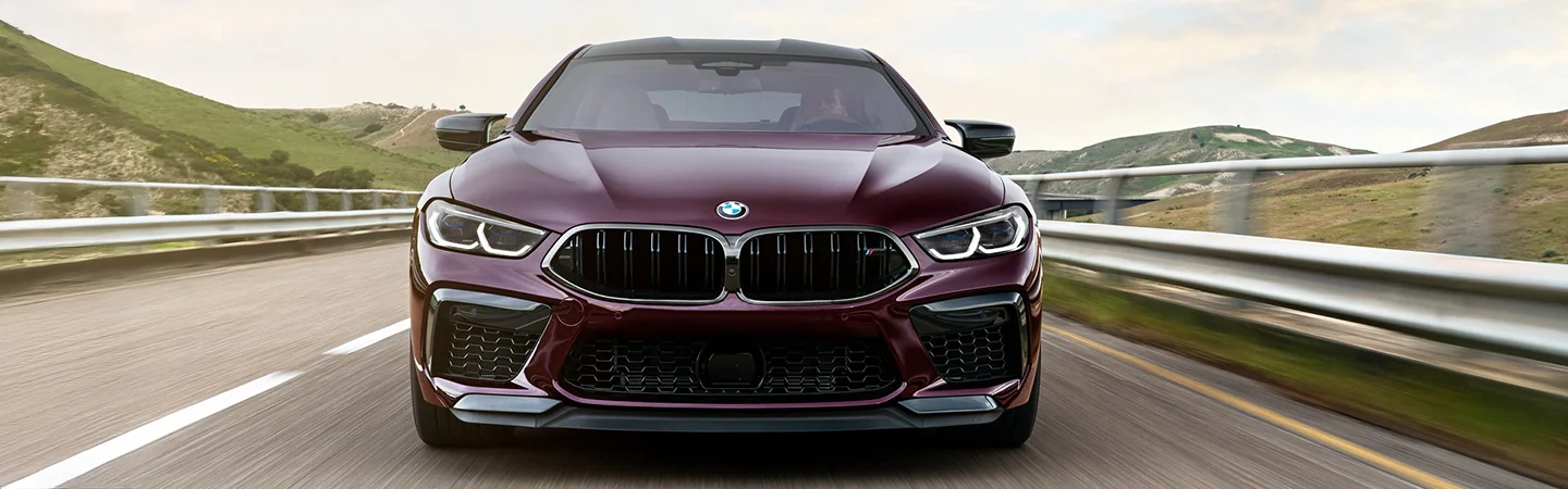 Front view of a 2025 BMW M8 Coupe driving on a road by hills