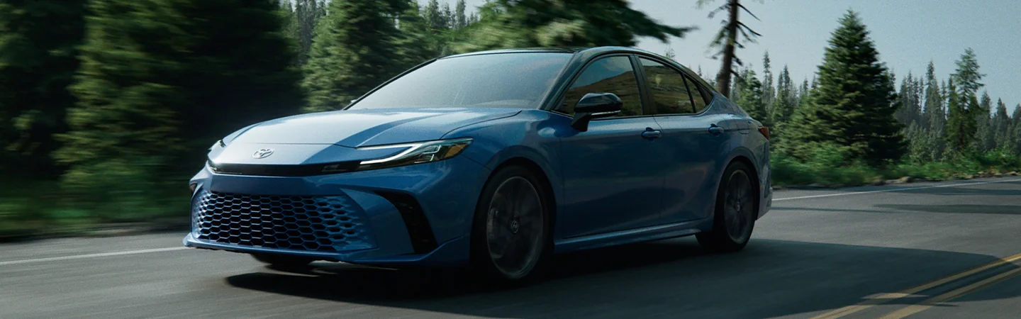 Front side view of a blue 2025 Toyota Camry driving on a road with a forest in the background