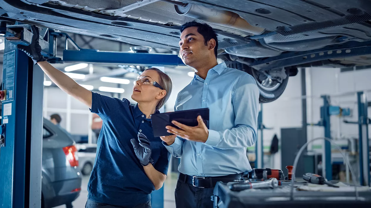 Mechanic under a car pointing things out to someone with a tablet