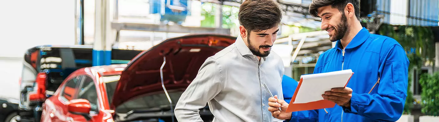 Two men examine a car parked in a garage, discussing its features and condition.