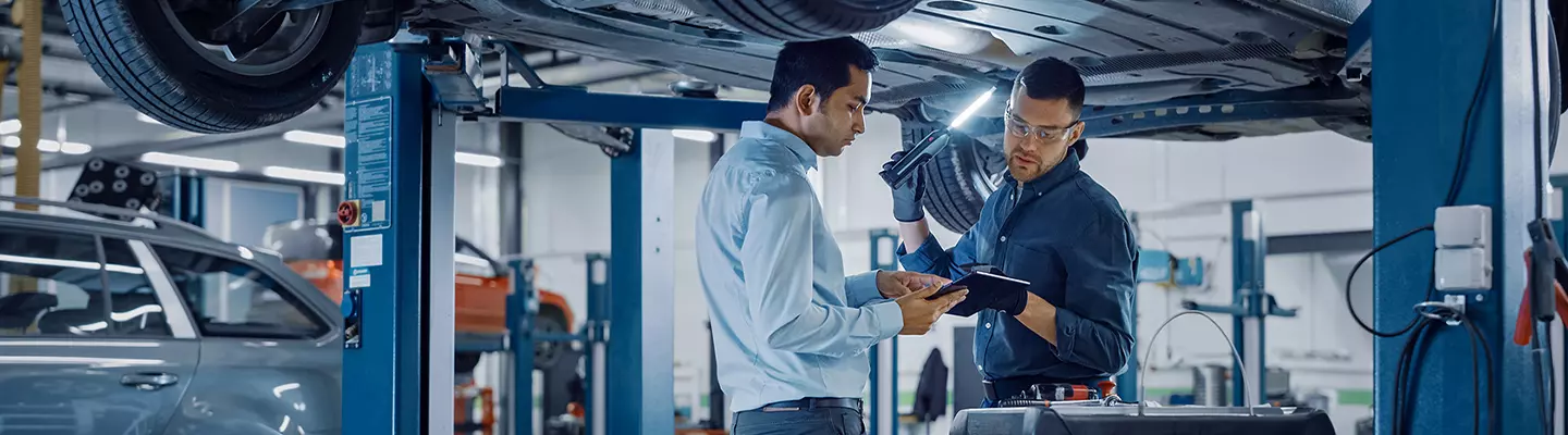 In a garage, two men are collaborating on car repairs, surrounded by tools and automotive parts.