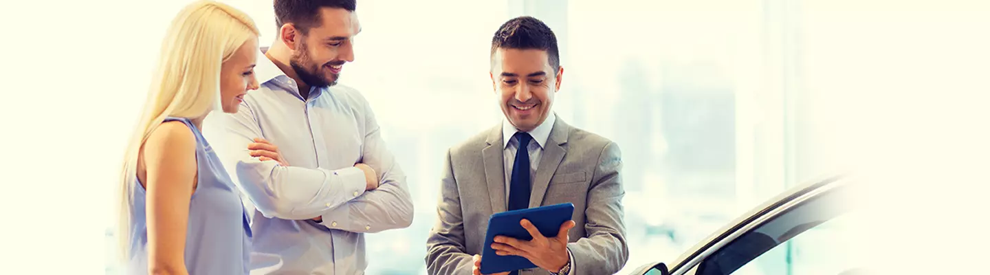 Three people gather around a car, one holding a tablet, discussing its features or specifications.