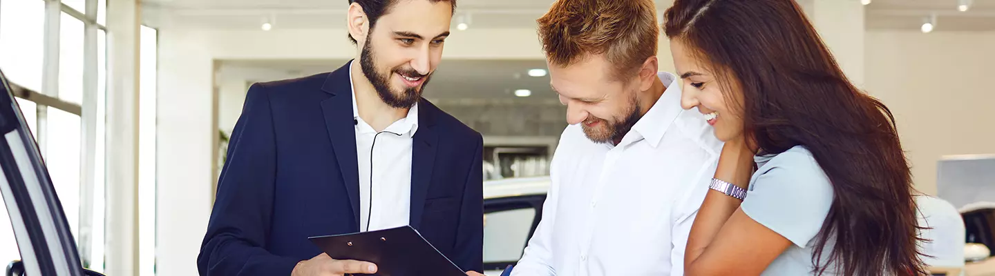 A couple looking at a car in a dealership, engaged in conversation about its specifications and pricing.