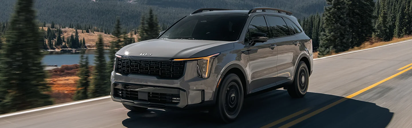 Front side view of a gray 2025 Kia Sorento driving on a road next to a lake and a forest
