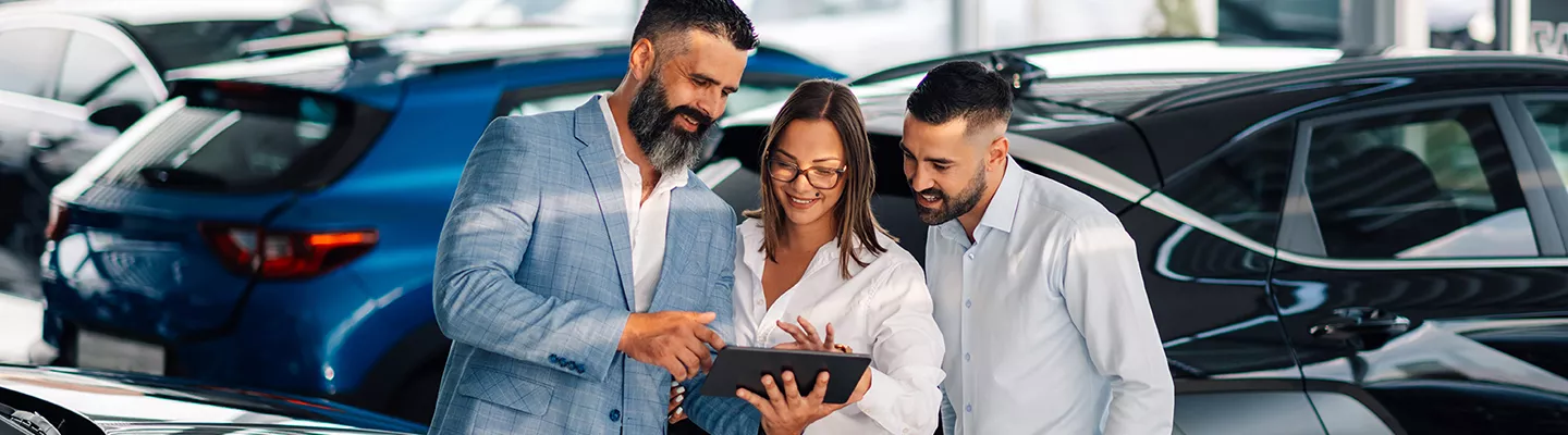Three people standing around a tablet smiling inside a car dealreship