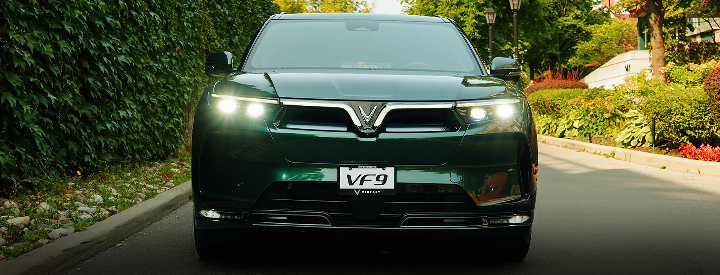 Front view of a green 2025 VinFast VF 9 driving by some houses