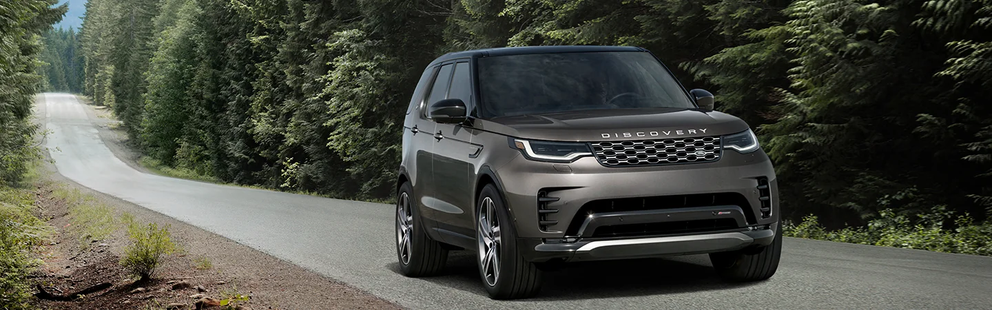 Front side view of a gray 2025 Discovery driving by some woods