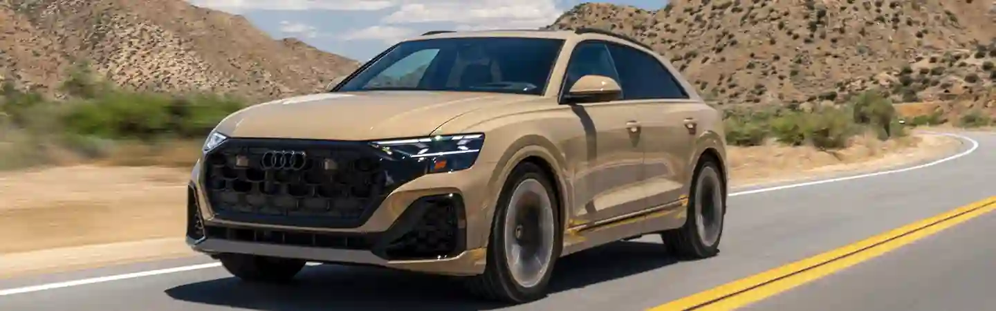 Front view of a tan 2025 Audi Q8 driving on a raod next to mountains