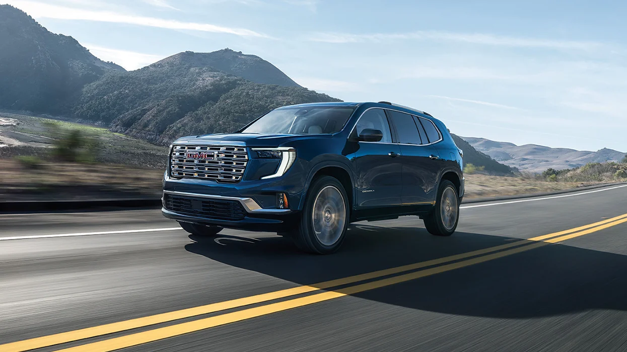 A blue 2026 GMC Acadia driving on a road next to some mountains