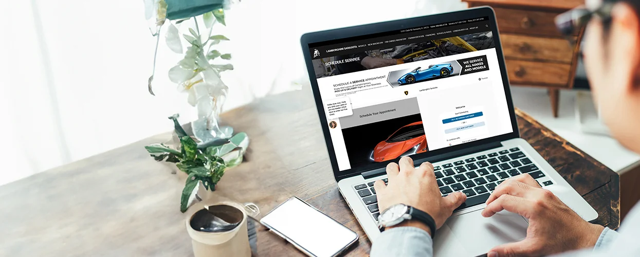 A man sits at a table, focused on a laptop screen displaying the Lamborghini of Sarasota website.