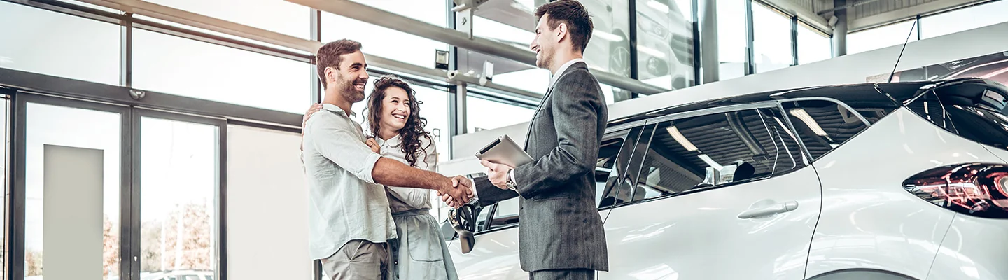 salesman shaking hands with husband and wife holding onto him while standing by a car