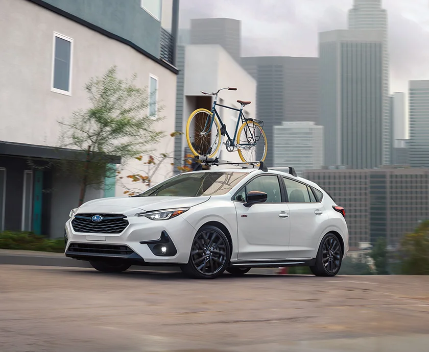 Front side view of a white 2026 Subaru Impreza driving in a city with bikes on the roof