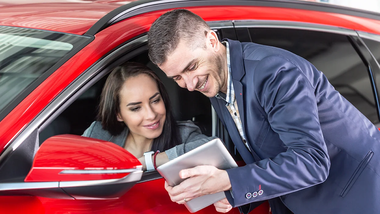 car salesperson standing speaking with customer in car's driver seat showing the advantages of hybrid car