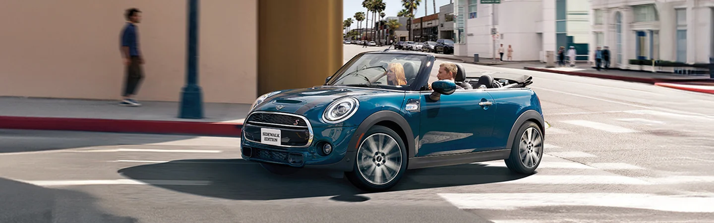 blue 2025 MINI Cooper Convertible with top down driving around a corner in city