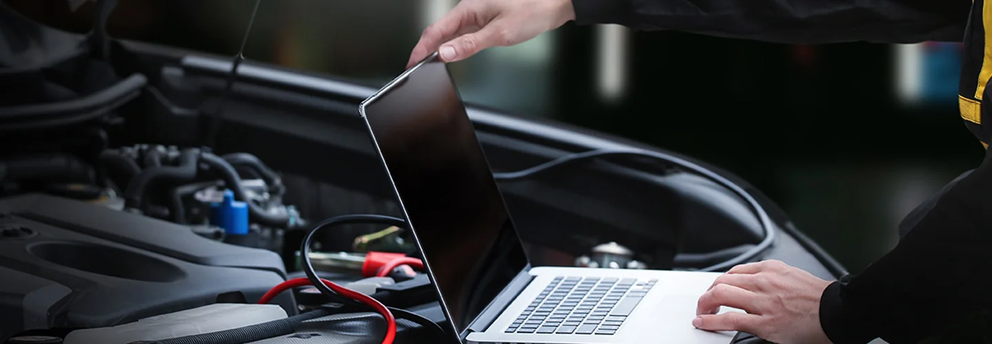 A man inspects his car's engine while using a laptop to gather information for troubleshooting.