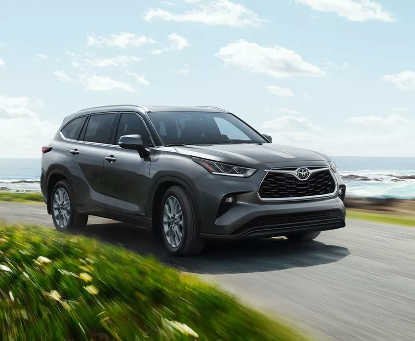 Front side view of a 2026 Toyota Highlander Hybrid driving on as road by the ocean in Tracy, CA